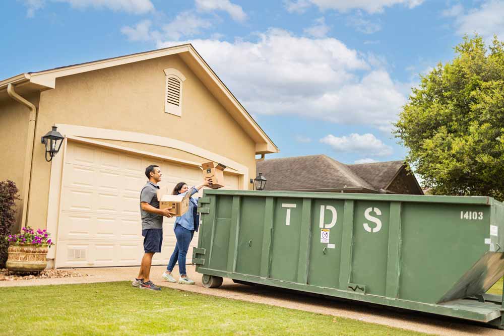 Green TDS mini roll off dumpster parked in front of house with couple throwing cardboard boxes into the dumpster for recycling