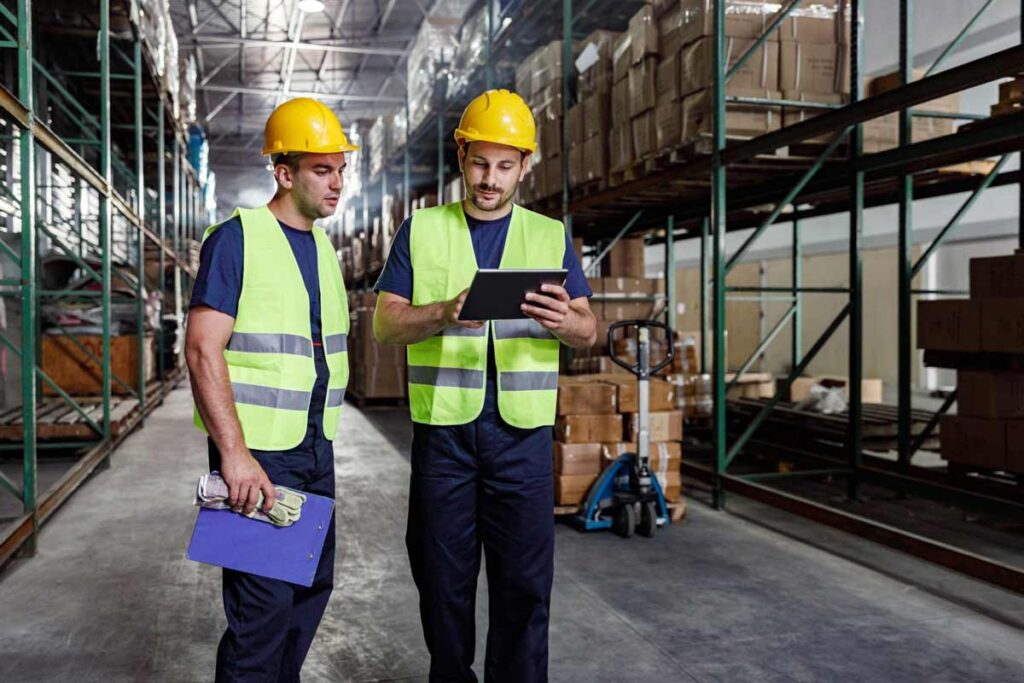 Two men working in a warehouse talking to each other and wearing hard hats