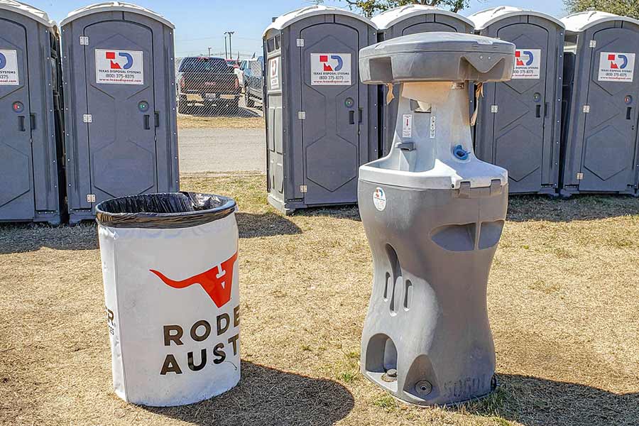 An Austin rodeo labeled waste container next to a gray hand washing station with portable restrooms in the background
